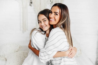 Anne ve kızı kameraya gülümsüyor ve beyaz oda arka planına sarılıyorlar. Mutlu bir aile..