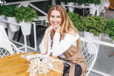 Güzel sarışın kadın kameraya gülümsüyor. Çiçekli bir kafedeki masada..