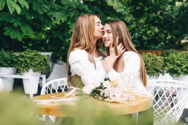 İki kız kardeşin el ele tutuşup kameraya gülümsediği bir fotoğraf. Biri elinde çiçeklerle.