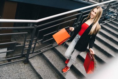 Mutlu genç kız elinde alışveriş torbalarıyla merdivenlerden aşağı iniyor. Şık kıyafet. Güzel sarışın. İndirim.