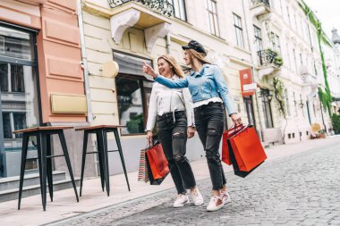 İki şık giyinmiş kız kardeşin alışveriş yaptıktan sonra caddelerde yürürken çekilmiş bir fotoğrafı. Biri mağazayı işaret ediyor..