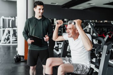 Spor salonundaki antrenör, sinsi bir adamla rehabilitasyon uygular, gülümser ve nasıl egzersiz yapılacağını söyler..