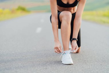 Uzun bir yolun arka planında koşmaya başlamadan önce ayakkabı bağı bağlayan bir kadının portresi.