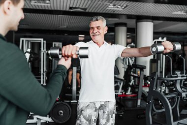 Rehabilitasyon. Kıdemli adam, eğitmenin gözetiminde bir salonda dambıllarla meşgul..