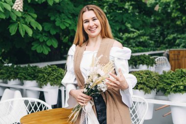 Elinde bir buket çiçek tutan ve kameraya gülümseyen genç bir kadının portresi. Açık hava fotoğrafı, bahar..