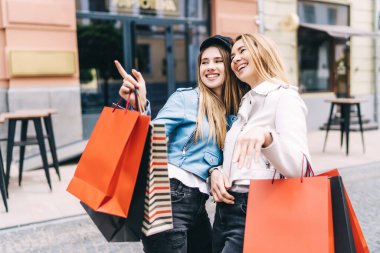 Caddenin ortasında alışveriş yaparken gülümseyen iki güzel kadın. Bir tanesi elini dükkanlara doğrultuyor..