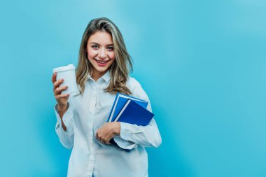 Mavi arka planda elinde kitaplar ve kahveyle genç bir kız öğrencinin portresi. Gülümseyerek kameraya bakıyor. Stüdyo fotoğrafı. Mesaj için yer. Arkaplan.