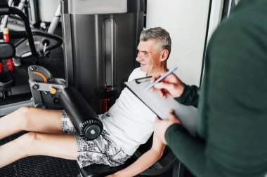 Gri saçlı bir adam spor salonundaki bir simülatör üzerinde egzersiz yapıyor, koçu sonuçları kaydediyor. Rehabilitasyon, eğitim planı.