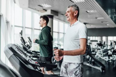 Spor salonunda koşu bandında iki adam, odak noktasında gri saçlı, gülümseyen son sınıf öğrencisi. Spor, eğitim, sağlık. Spor Salonu.