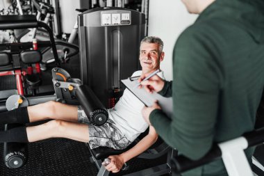 Konferansta simülatör üzerinde alıştırma yapan kıdemli adam, antrenörü eğitim planını ve sonuçları yazıyor. Rehabilitasyon koçu.