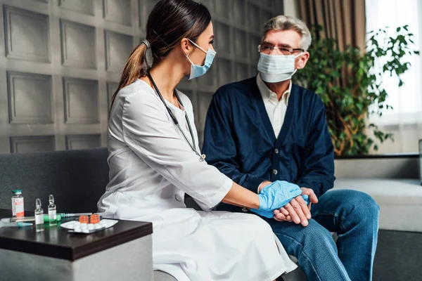 A nurse in a protective mask and gloves holds the hand of an older man in a protective mask, supporting and helping the elderly. Care for the elderly. Doctor\'s visit home. Pandemic, covid-19.