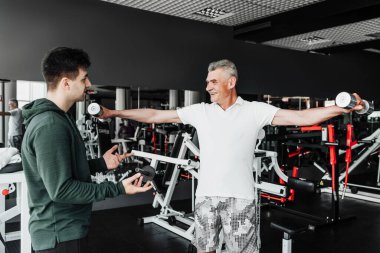 Spor salonunun ortasında, emekli bir adam dambıllarıyla egzersiz yapıyor, eğitmeni onu izliyor ve destekliyor. Spor, spor, sağlık.