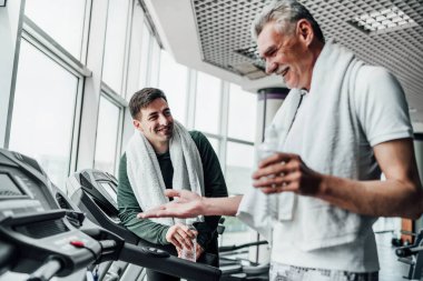 Spor salonundaki iki adamın portresi, koşu bandında birbirleriyle iletişim kurup su içiyorlar. Kardiyo, spor, sağlık.