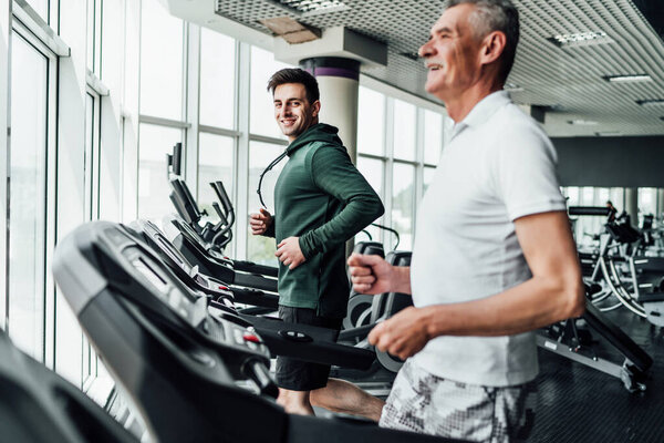 Two people on the treadmill, in the focus of a handsome young coach, he is smiling at the camera. Sports, gym, healthy lifestyle.