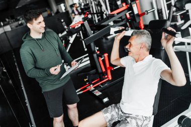 İki adam spor salonunda, biri elinde eğitim planı tutan bir fizyoterapist, diğeri de simülatör üzerinde egzersiz yapıyor..