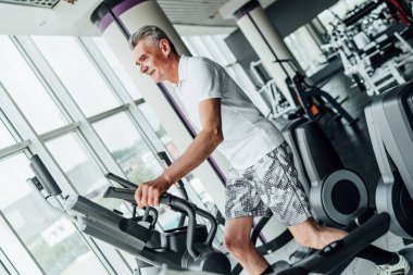 Yaşlı bir adamın portresi bir kardiyo makinesinde egzersiz yapıyor, önünde büyük panoramik pencereler, modern geniş bir spor salonunda..