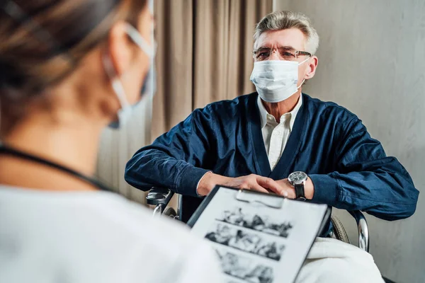 Portrait of a senior man with a protective mask and in a wheelchair listening to a doctor. Doctor\'s visit to the patient, treatment.