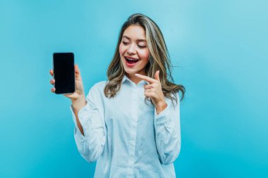 Ne mutlu ki küçük kız telefonu siyah ekranla işaret ediyor. Mavi arka planda. Stüdyo fotoğrafı. Reklamcılık. Ticaret, online mağaza.