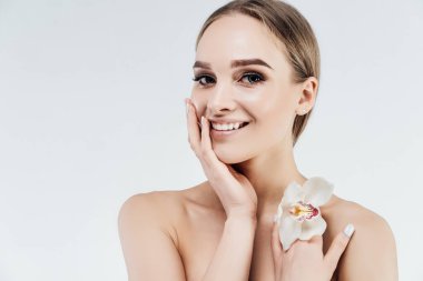 Beyaz bir arka planda beyaz tenli çekici ve nazik bir kız. Güzel ve temiz bir teni var. Elini yüzüne dayayıp kameraya gülümsüyor..