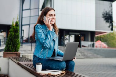 Elinde bilgisayarla çekici bir kadın sokakta bir bankta oturuyor, serbest çalışıyor, ofis merkezi, internet eğitimi, uzaktan çalışıyor..