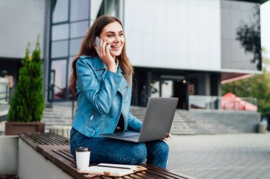 Çekici bir kız telefonda konuşuyor ve sokakta bir bilgisayar üzerinde çalışıyor bir bankta oturuyor, ofis merkezinin arkasında, yaşam tarzı, serbest çalışma.