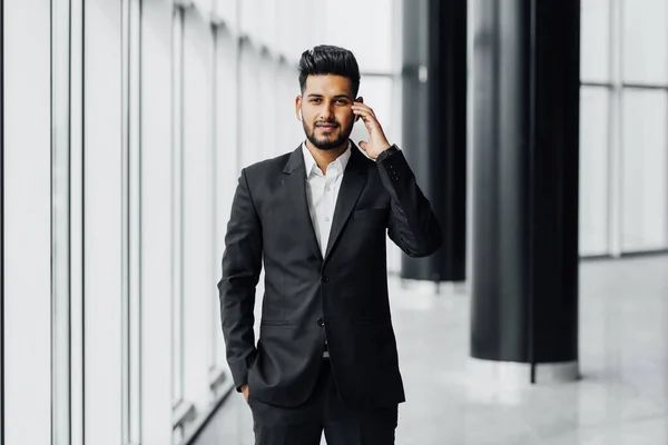 An attractive bearded Indian businessman in a modern office center, wearing a black suit, holds his hand near a wireless earphone and looks at the camera. Young Indian businessman.