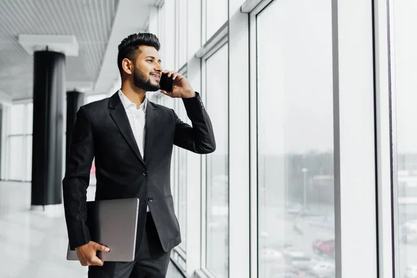 Indian serious smiling businessman uses his phone, working negotiations, holding his computer by the window in the office. Young Indian businessman. Backgraund.