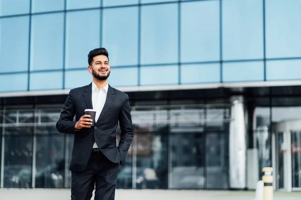 Modern bearded Indian man, smiling goes to a business meeting, behind him a large office building, a successful Indian businessman.