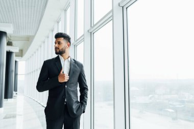 Modern bir binada, büyük bir pencerenin yanında siyah takım elbiseli modern bir Hintli adam, ofis çalışanı, banka çalışanı..