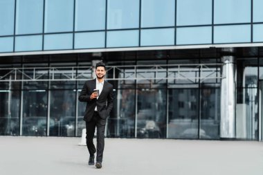 Siyah takım elbiseli Hintli bir adam iş görüşmesi için caddede yürüyor, arkasında büyük, modern bir bina var..