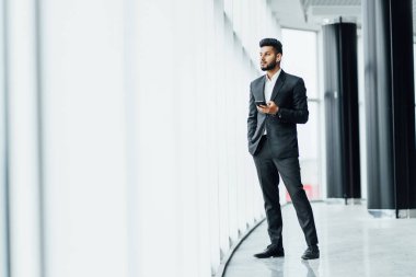 Yakışıklı, genç, sakallı bir Hintli pencerenin yanındaki modern bir binanın ortasında duruyor. Siyah bir takım elbise giymiş, bir ofis çalışanı..