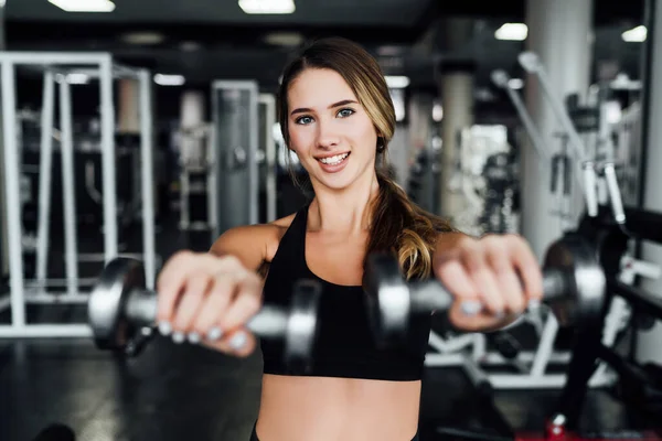 Beautiful sports girl in the gym reaches out with dumbbells to the camera, she smiles and looks at the camera, lifestyle.