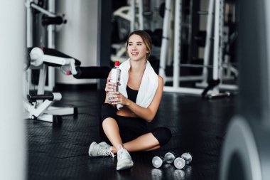 Genç ve çekici bir kız modern bir spor salonunda yerde dinleniyor ve su içiyor..