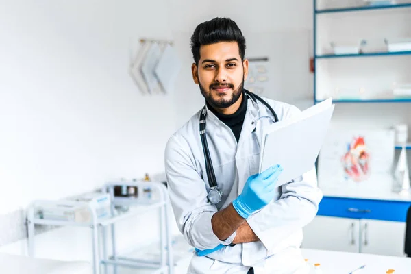 A handsome Indian doctor man in a medical suit in the middle of a modern hospital, he is looking at the camera.