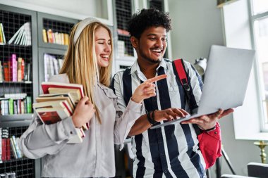 Mutlu öğrenciler konuşarak ve bilgisayara bakarak eğleniyorlar, kitaplarla dolu bir rafın arkasında..
