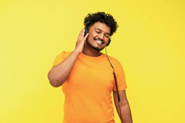 Neşeli gülümseyen Hintli adam sarı arka planda izole edilmiş kulaklıklar, modern cihazlar, sarı arka planda mesaj, stüdyo fotoğrafı için yer var..