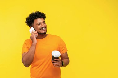 Sarı arka planda izole edilmiş Hintli bir öğrenci. Mesajlaşmak için yeri var. Akıllı telefonu elinde tutuyor, gülümsüyor..