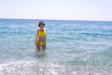 Yaz tatili tarzı, kumsalda yürüyen sarı bikinili, ince, kızıl saçlı güzel bir kız. Türkiye 'de mavi kristal berrak denizde mutlu gezgin kadınlar, Kemer.