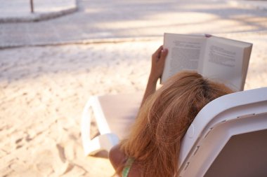 Güzel genç bir bayan havuz kenarında uzanıyor ve kitap okuyor. Güverte sandalyesinde rahatça kitap okuyan bir kadın. Yaz tatili