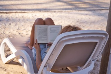 Güzel genç bir bayan havuz kenarında uzanıyor ve kitap okuyor. Güverte sandalyesinde rahatça kitap okuyan bir kadın. Yaz tatili