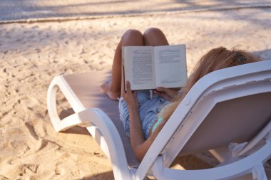 Güzel genç bir bayan havuz kenarında uzanıyor ve kitap okuyor. Güverte sandalyesinde rahatça kitap okuyan bir kadın. Yaz tatili