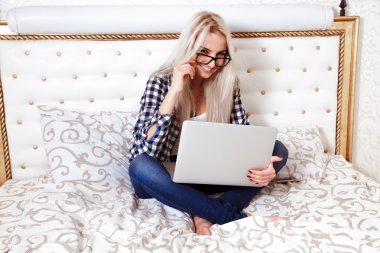 Young blonde woman with beautiful smile sits on bed with laptop