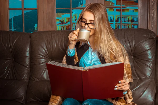 A girl sits at a window on the couch, reading a book and drinkin