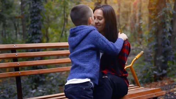 若いです未成年の子供キス彼の幸せな母親で彼女の鼻で彼女のベンチにザ公園で暖かい秋の日 — ストック動画