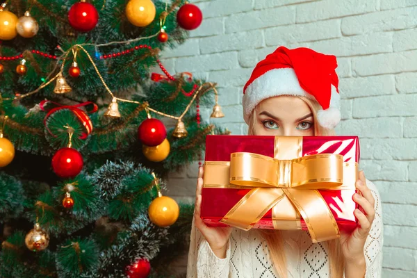 Charmante Blondine mit Weihnachtsmütze versteckt ihr Gesicht hinter einem großen Weihnachtsgeschenk — Stockfoto