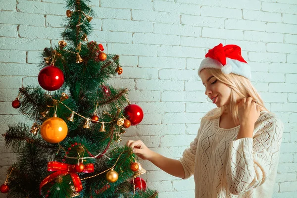 Веселая молодая блондинка украшает рождественскую елку в шляпе Санты — стоковое фото