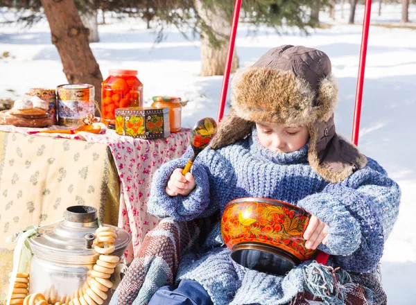 Malý kluk v čepici s krytkami na uši hraje winter park. Ruský styl na pozadí samovar a bagely — Stock fotografie