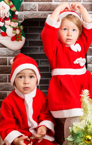 Broer en zus gekleed Santa Claus kostuum door open haard. Kerstmis en Nieuwjaar — Stockfoto