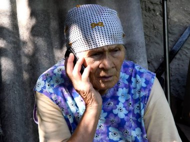 80 yaşında, geleneksel giysiler içinde cep telefonuyla konuşan bir kadın. Kırsal kesimde, kafası mendile sarılı, eli buruşmuş akıllı telefonlu yaşlı bir kadın.
