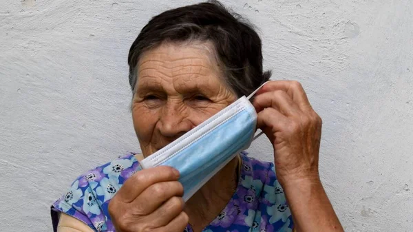 Elderly wrinkled woman putting on protective medical face mask. Old lady shows how to wear mask in appropriate way. Senior people protection while lockdown